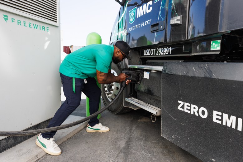 A person wearing a black hat, green shirt and navy blue pants inserts a charging nozzle into the side of a black and green commercial truck.