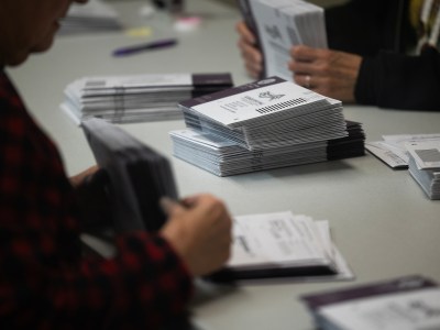 Stacks of purple and white mail-in ballot envelopes sit on a stable as election workers sort them. The arms and hands of the workers are visible, but the faces are not. The focus is on the ballots.