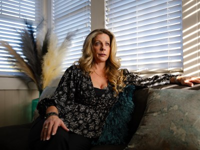 A person with long, wavy blonde hair sits on a couch, leaning back with one arm resting on the armrest. They are wearing a black, floral-patterned blouse and dark pants. Sunlight streams through window blinds, casting soft shadows across the room. Decorative pampas grass and textured pillows are visible in the background, adding to the cozy indoor setting. The person gazes into the distance with a serious expression.