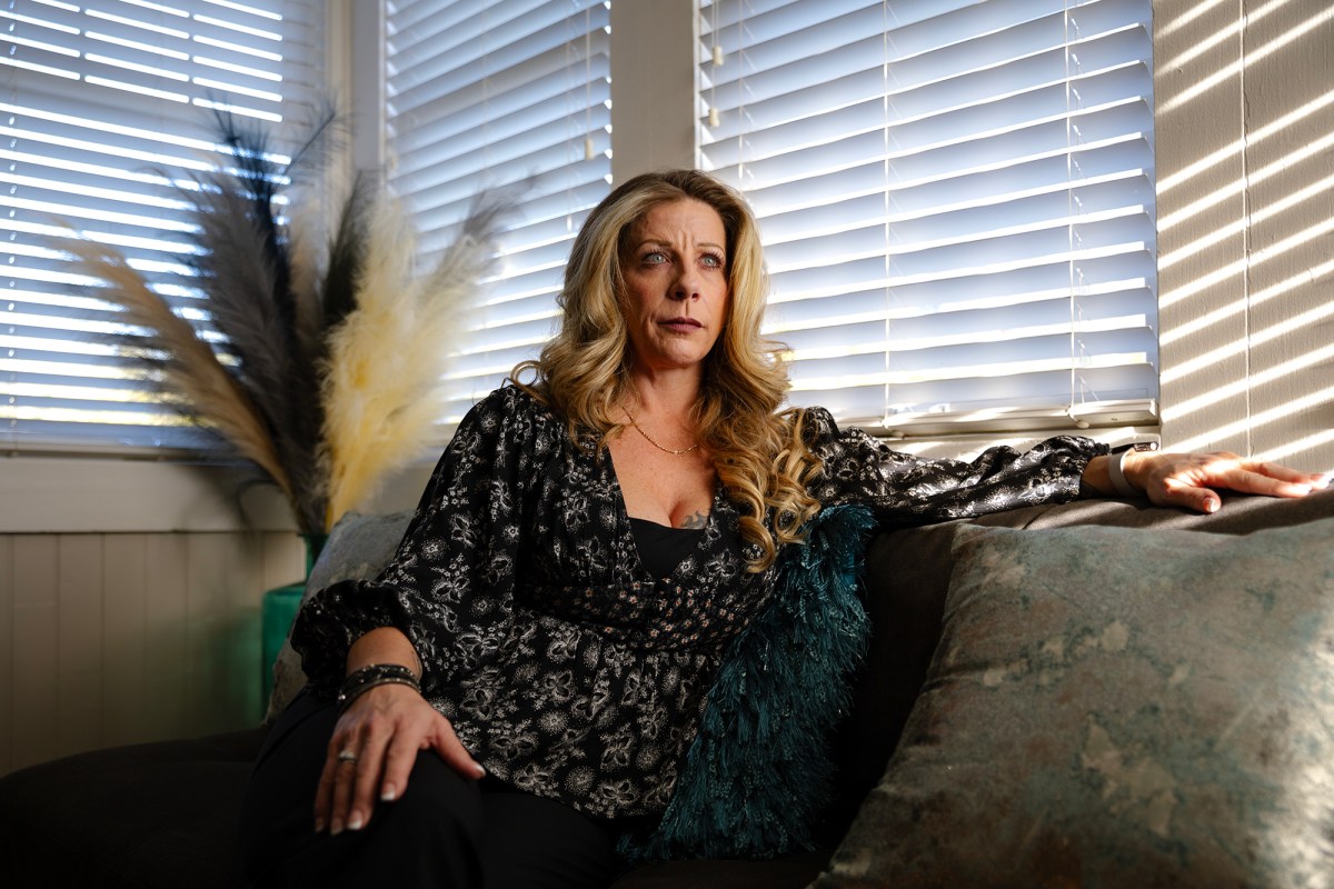 A person with long, wavy blonde hair sits on a couch, leaning back with one arm resting on the armrest. They are wearing a black, floral-patterned blouse and dark pants. Sunlight streams through window blinds, casting soft shadows across the room. Decorative pampas grass and textured pillows are visible in the background, adding to the cozy indoor setting. The person gazes into the distance with a serious expression.