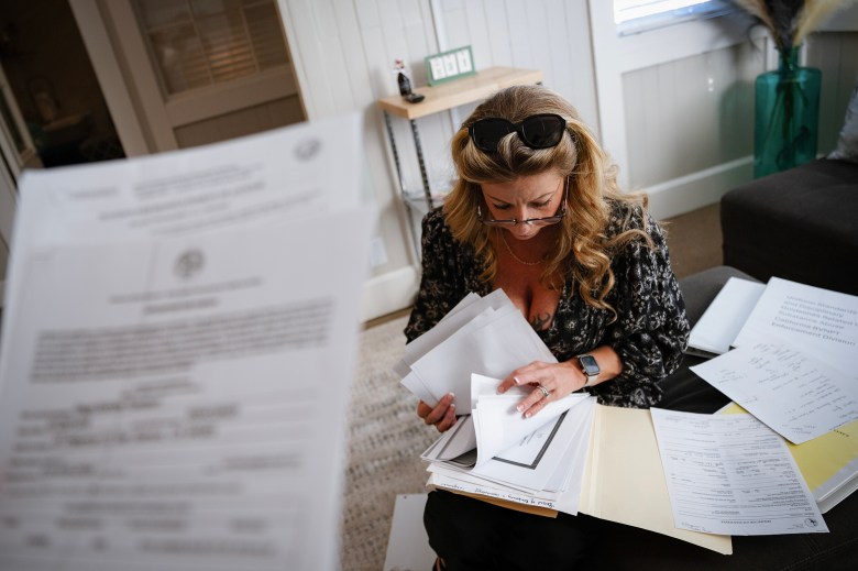 A person with long, wavy hair sits on the floor surrounded by papers, holding and organizing a stack of documents. They are wearing a patterned blouse, glasses on their face, and sunglasses resting on their head. A large folder and various papers, some with writing and printed text, are spread across the area, creating a cluttered workspace. The person is focused on reviewing the paperwork.