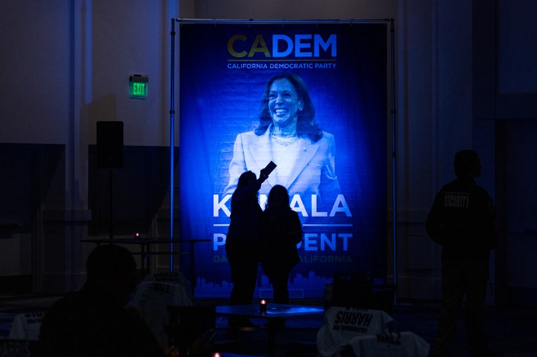 The silhouette of two people taking a selfie with their phone in front of a "Kamala For President" banner with Harris' photo at a watch party in Los Angeles.