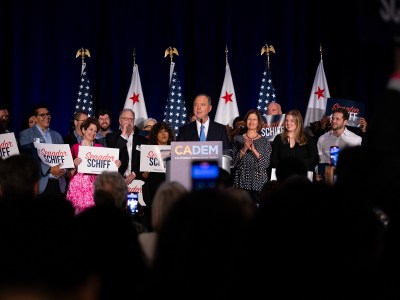 A person in a suit stands at a podium. A sign on the podium reads "CA DEM" and "California Democratic Party." People stand in the background while holding signs that read "Senador Schiff." California and U.S. flags stand behind the group.