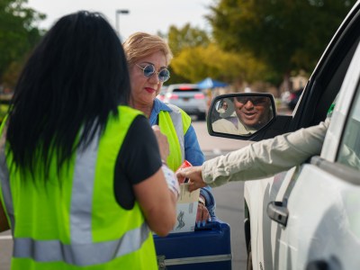 A person driving a car extends their hand out the window to insert their ballot envelope into a drop box being held by an election worker. The back of another election worker can be seen nearby.