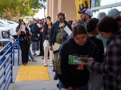 People wait in line on the sidewalk of a light brown building during election day. Some people hold their ballot envelops in their hands.