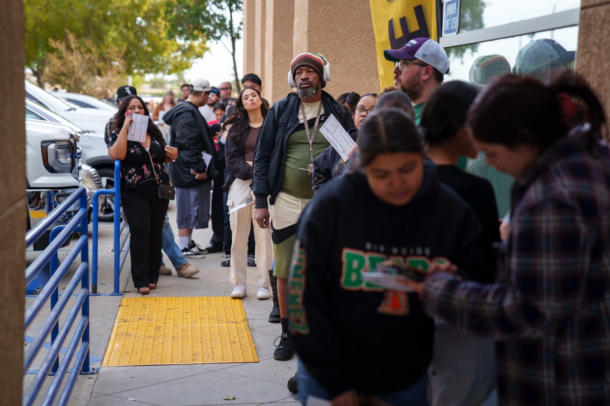 People wait in line on the sidewalk of a light brown building during election day. Some people hold their ballot envelops in their hands.