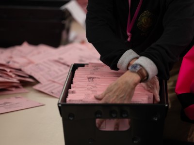 A close-up view of a person's hands sifting through a small black basket filled with pink mail-in ballot envelopes. A stack of pink mail-in ballot envelopes can be seen laying on the table in the background.