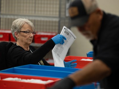 A person wearing blue medical gloves lifts up a ballot while they sit in front of a table with blue and red boxes filled with voter ballots. Another person working nearby can be seen in the foreground.