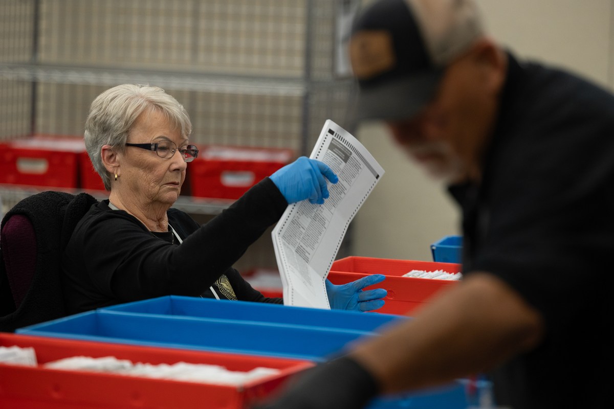 A person wearing blue medical gloves lifts up a ballot while they sit in front of a table with blue and red boxes filled with voter ballots. Another person working nearby can be seen in the foreground.