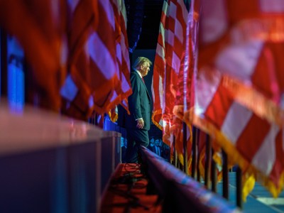 A side profile of a person in a suit and red tie walking amidst large, partially blurred flags in a dimly lit setting. The scene has a dramatic, almost solemn ambiance, with vivid red and blue lighting casting reflections on the surrounding flags, creating a sense of depth and movement.