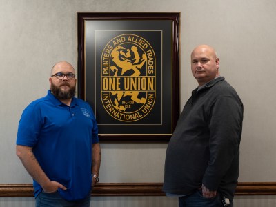 Two people stand in front of a framed International Union of Painters and Allied Trades logo hung up on a wall in an office. The person on the left