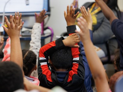 A student wearing a black and red sweater raises their hand among other students also raising their hands during class.