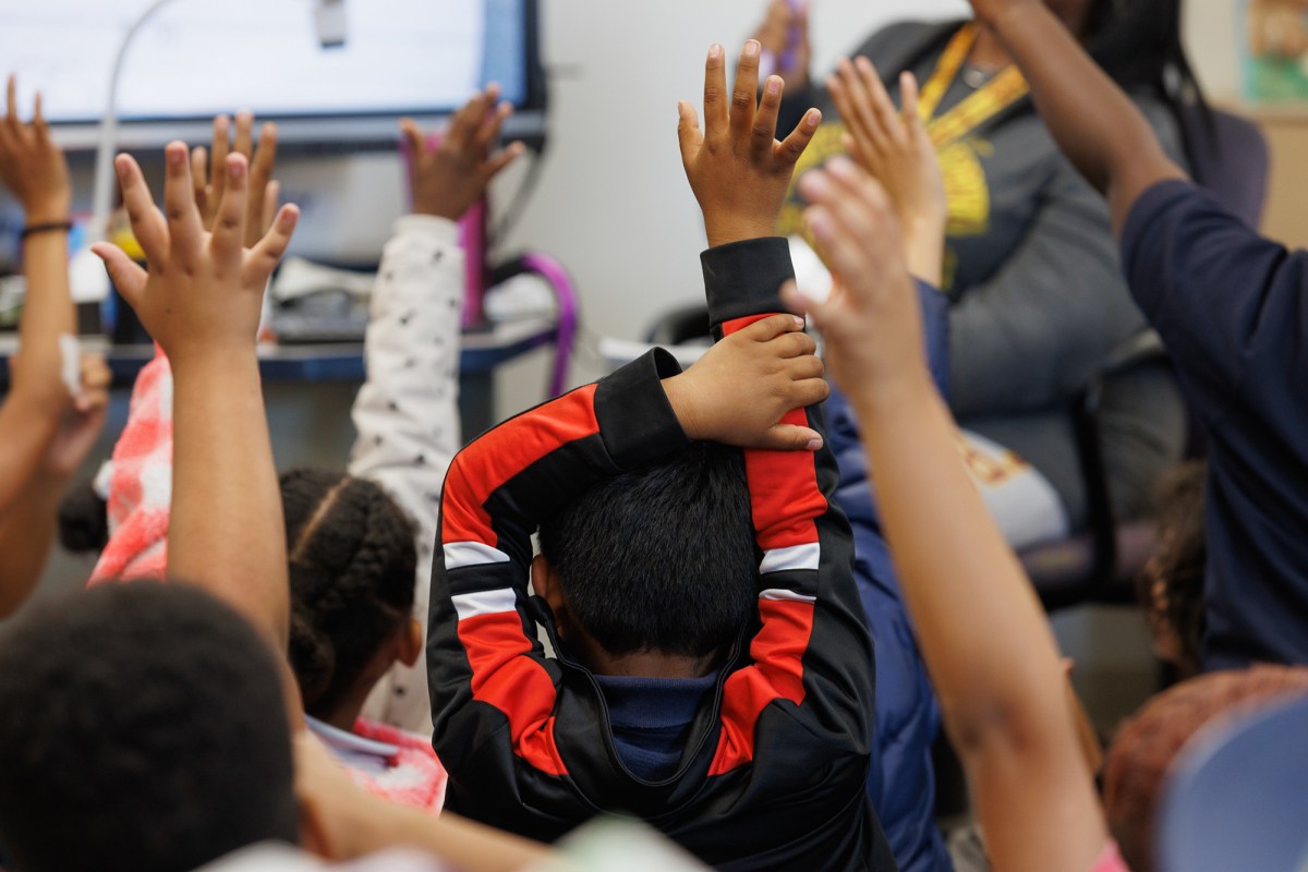 A student wearing a black and red sweater raises their hand among other students also raising their hands during class.