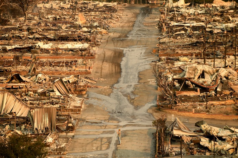 A person stands in the middle of a road surrounded by charred home structures that were destroyed by a wildfire.