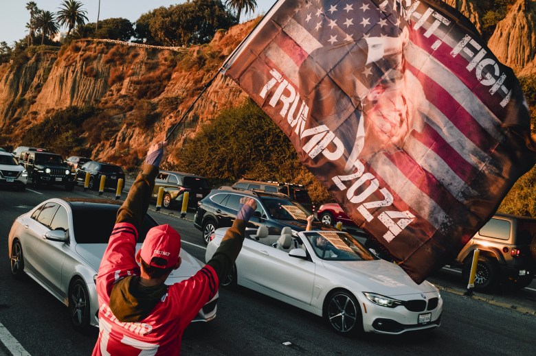 A person wearing a red 'Trump' hat and jersey holds a large flag that reads 'Trump 2024' with an image of Donald Trump over a U.S. flag backdrop. They face a line of cars, some with passengers who have their arms raised, as they drive along a coastal road bordered by rugged cliffs and tall palm trees, illuminated by warm sunlight.