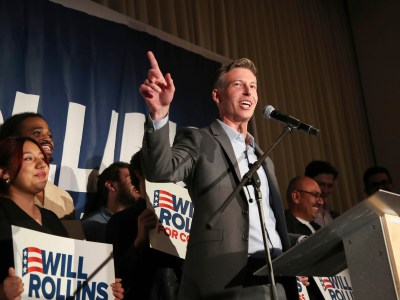 A person points as they speak at a microphone at a podium with people behind him holding up signs.