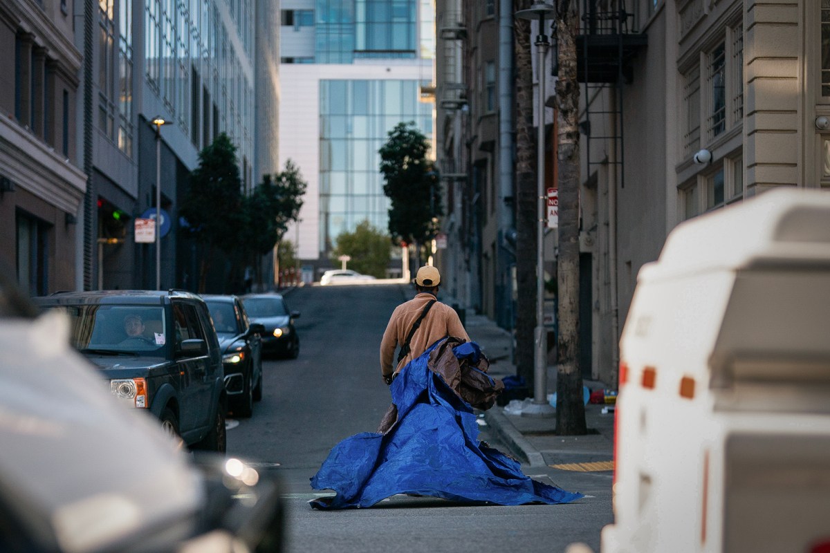 A person wearing a beige jacket and cap walks down a city street, pulling a large, crumpled blue tarp. The scene is framed by tall buildings, parked cars, and a modern glass structure in the background. The muted urban setting is illuminated by soft, natural light, highlighting the quiet and solitary moment.