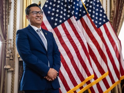 A person wearing glasses and dressed in a blue suit stands with his hands crossed in front of a row of American flags in a decorated office.