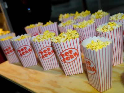 White popcorn boxes with red pinstripes, and the words "POP CORN" printed on them sit on a light colored, wooden table. The setting is a theatre or movie event.