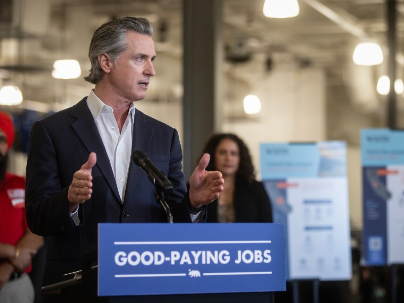 A person in a suit speaks at a podium labeled "GOOD-PAYING JOBS," gesturing with their hands. Behind them, two individuals stand near informational boards with text and graphics. The setting appears to be an indoor event space with industrial lighting.