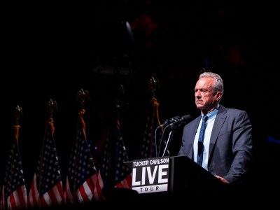 A person stands on stage at a podium with microphones next to a row of American flags.