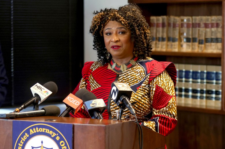 A person wearing a red and gold pattern dress stands behind a lectern with microphones from local media outlets during a press conference. A bookshelf with law books can be seen behind them.