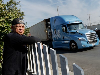 A person wearing a black backwards hat and shirt leans over the white fence as a blue and white commercial truck drives down a street on a sunny day.