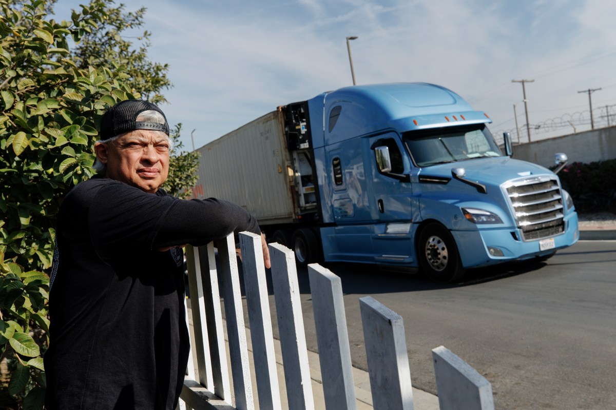 A person wearing a black backwards hat and shirt leans over the white fence as a blue and white commercial truck drives down a street on a sunny day.