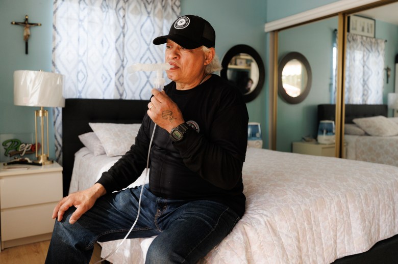 A person wearing black hat and shirt with blue jeans sits on the edge of a full size bed with white bedding as they breath into a tube connected to medical vaporizer.