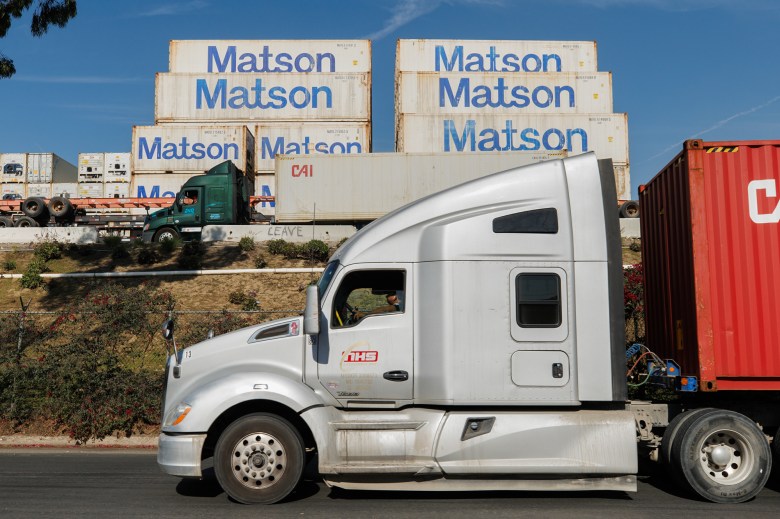 A gray and white commercial truck drives down a street. Another green and white commercial truck can be seen up a hill in the distance alongside several cargo containers labeled "Matson" on the side.