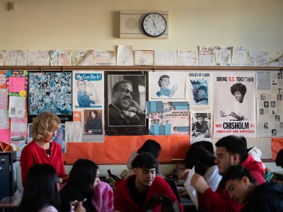 A wide view of various posters related to democracy and prominent figures hung up in a classroom wall. Notable figures include U.S. history including Activist Malcom X, Supreme Court Justice Sonia Sotomayor, Activist Cesar Chavez and Congresswoman Shirley Chisholm. A group of students and teacher can be seen sitting on desks in the foreground.