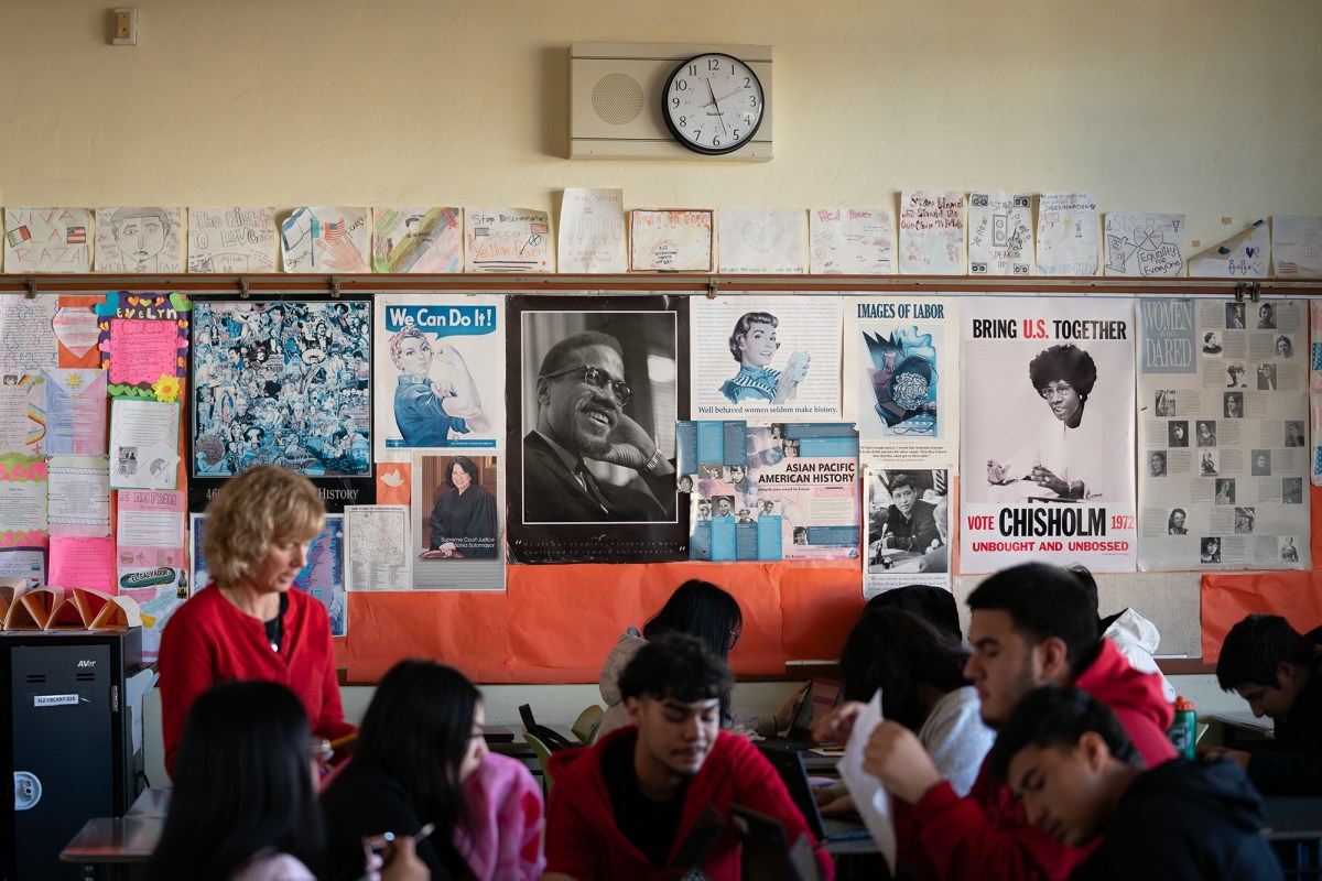 A wide view of various posters related to democracy and prominent figures hung up in a classroom wall. Notable figures include U.S. history including Activist Malcom X, Supreme Court Justice Sonia Sotomayor, Activist Cesar Chavez and Congresswoman Shirley Chisholm. A group of students and teacher can be seen sitting on desks in the foreground.