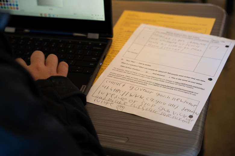 A close-up view of a student's paperwork next to them as they type on a laptop. The paper contains notes on how to contact California Gov. Gavin Newsom.