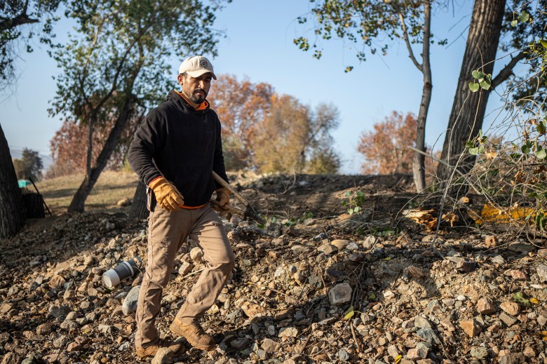 A person in a working sweatshirt and protective work attire walks through rough terrain in a park with pruning shears under their arm.