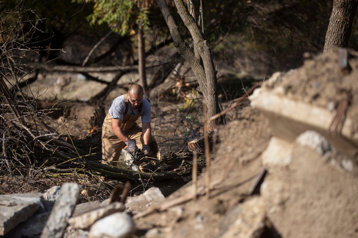 A person in working overalls uses a chainsaw to cut into a tree trunk in a park. Debris from concrete slabs and a chopped-down tree surround the person as they work in a park.