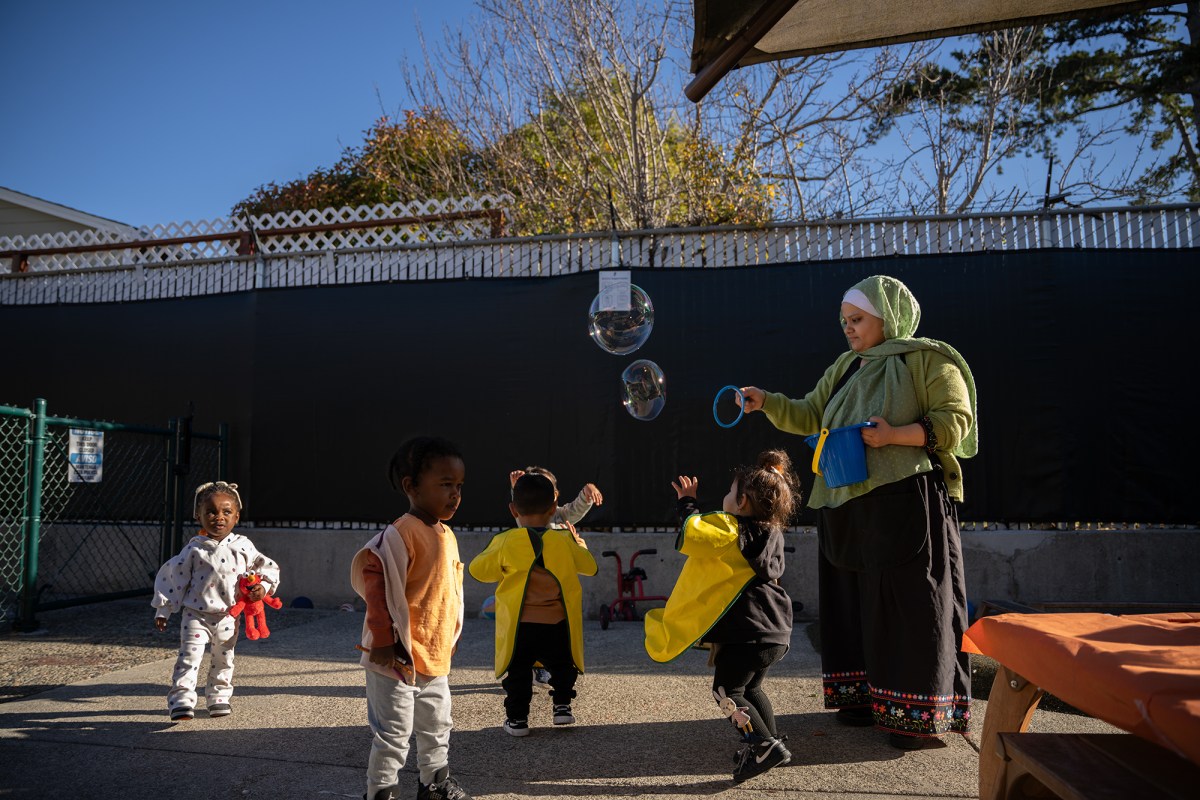 A person, wearing a green hijab and blouse with a long black skirt, uses a bucket and a ring device to release bubbles to a group of children playing nearby.