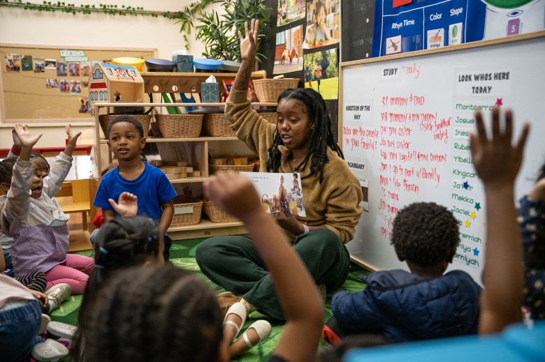 A teacher, wearing a fluffy light brown sweater abd green pants, raises their hand and holds a book while they sit on the ground surrounded by young children with their hands raised.