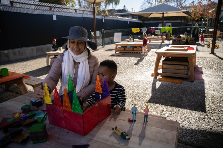 A person wearing a grey hat, white scraf and a pink thermal jacket, uses magnetic shapes to build a castle with a young child next to them.