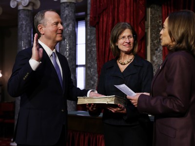 A lawmaker, standing on the left and wearing a black suit with a purple tie, holds their right hand up while placing their left hand on a book. A person, standing in the center and wearing a black blazer, holds the book as they look towards Vice President Kamala Harris, standing on the right and wearing a purple blazer, as she directs the swearing-in ceremony.