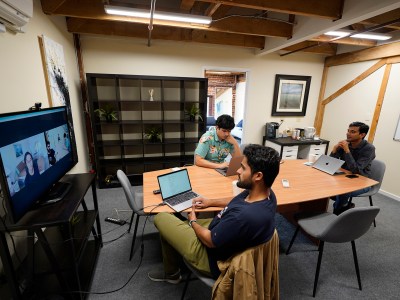 Three people sit around a table in an office meeting room with laptops in front of them. One person is looking at their computer while the other two look at a television screen with two people other people in a video meeting call.