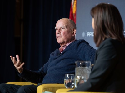 A side view of elderly person wearing a blue knitted sweater sitting in a yellow chair on stage next to another person during an event.