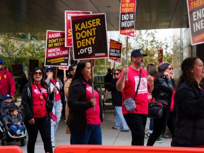 A group of people in front of a building holding picket signs that reads "kaiserdondeny.org" and "stand with kaiser therapist, end the inequity."