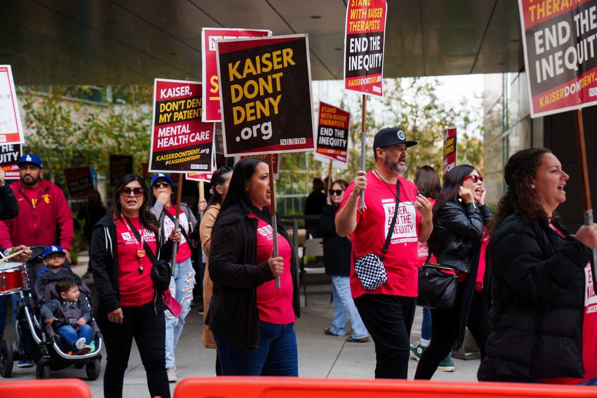 A group of people in front of a building holding picket signs that reads "kaiserdondeny.org" and "stand with kaiser therapist, end the inequity."