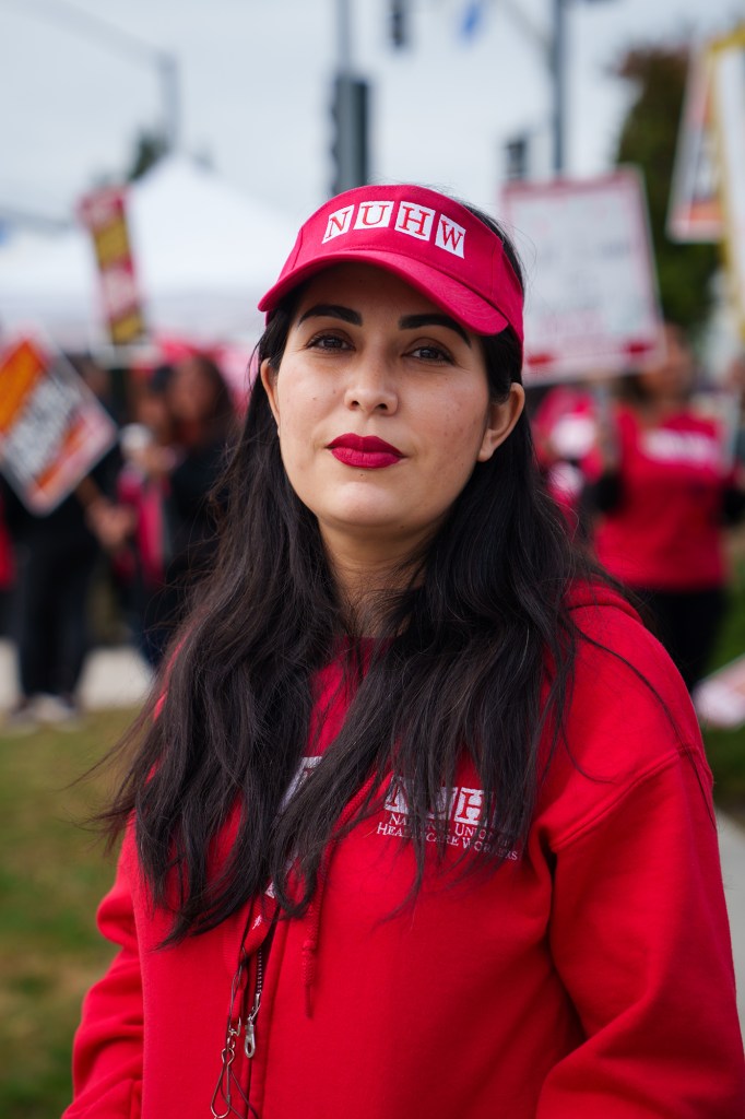 A portrait of a woman with long black hair wearing a red and sweater hat with the "NUHW" logo on it during a strike.