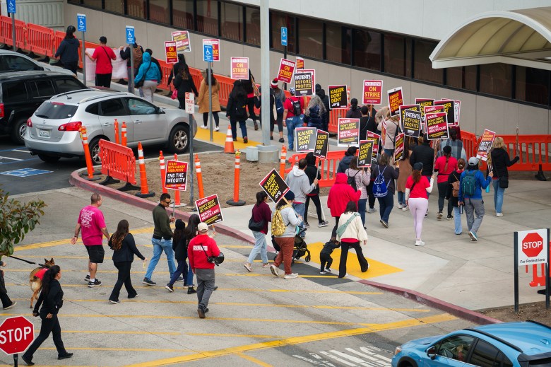 A wide view of a group of people in front of a building holding picket signs that reads "kaiserdondeny.org" and "stand with kaiser therapist, end the inequity."