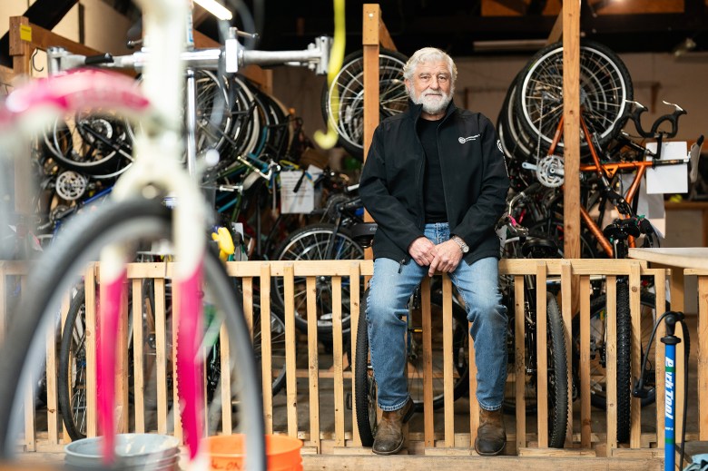 A person wearing a black jacket is leaning against a wooden rail with bicycle tires in the slots. Behind the person are sets of bicycle racks with bikes hanging inside a repair workshop.