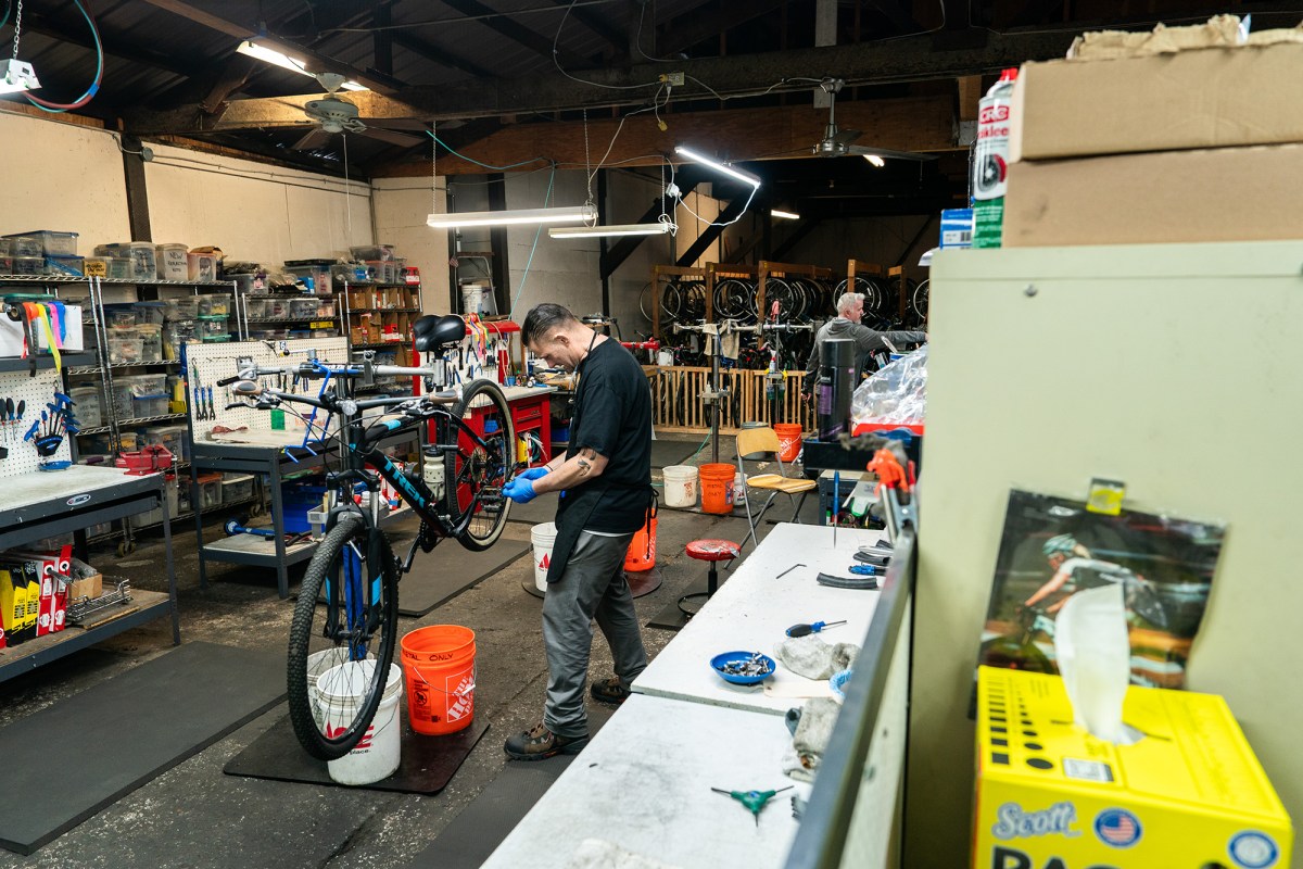 A person wearing blue gloves working on the back tire of a bicycle that is being held up in the center of a workshop. The workshop has buckets on the ground and tables with different tools around the edges, and bike racks with bicycles in the background.