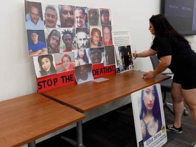 A person arranges posters of people who have died while in-custody in San Diego jails on a table during an event. One poster, containing photos of 19 individuals, reads "stop the deaths!" at the bottom.