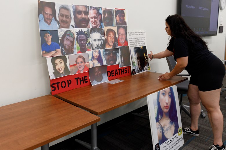 A person arranges posters of people who have died while in-custody in San Diego jails on a table during an event. One poster, containing photos of 19 individuals, reads "stop the deaths!" at the bottom.