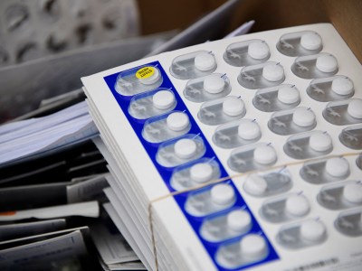 A stack of small white plastic containers with white pills inside plastic bubbles with one with a yellow sticker saying "Start Here". The stack is held with a rubber band and sits on a desk on a stack of papers.